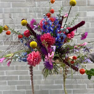 Unser Signature-Strauß feiert die schönste Zeit des Jahres mit einer Explosion aus Farben und Texturen. Im Mittelpunkt stehen prächtige Dahlien in kräftigem Pink und Fuchsia, die Romantik und Eleganz versprühen. Flankiert werden sie von den majestätischen, tiefblauen Blütenkerzen des Rittersporns, die dem Arrangement eine edle Tiefe und eine aufregende Höhe verleihen. Das Besondere liegt im Detail: Knallgelbe, kugelrunde Trommelstöckchen (Craspedia) setzen moderne, grafische Akzente, während die leuchtend orangefarbenen Lampions der Physalis und die samtig herabhängenden Rispen des Fuchsschwanzes (Amaranthus) dem Strauß einen charmanten, herbstlichen Touch verleihen. Feine Ziergräser und filigrane Beiblüten runden das Meisterwerk ab und lassen den Strauß wirken wie frisch auf einer Sommerwiese gepflückt.